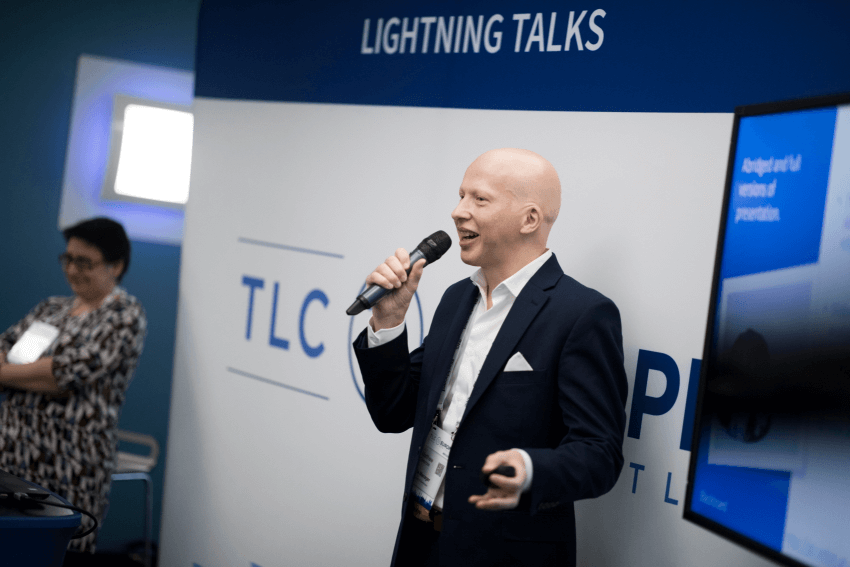 Matthew Deeprose, speaking at the Blackboard European Teaching and Learning Conference. Matt is wearing a blue suit with a white shirt and white pocket square. Holding a microphone, Matt is enthusing his audience.